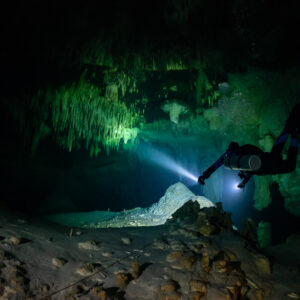 Sidemount diver lighting a rocky cave floor with stalactites above, Quintana Roo, Mexico