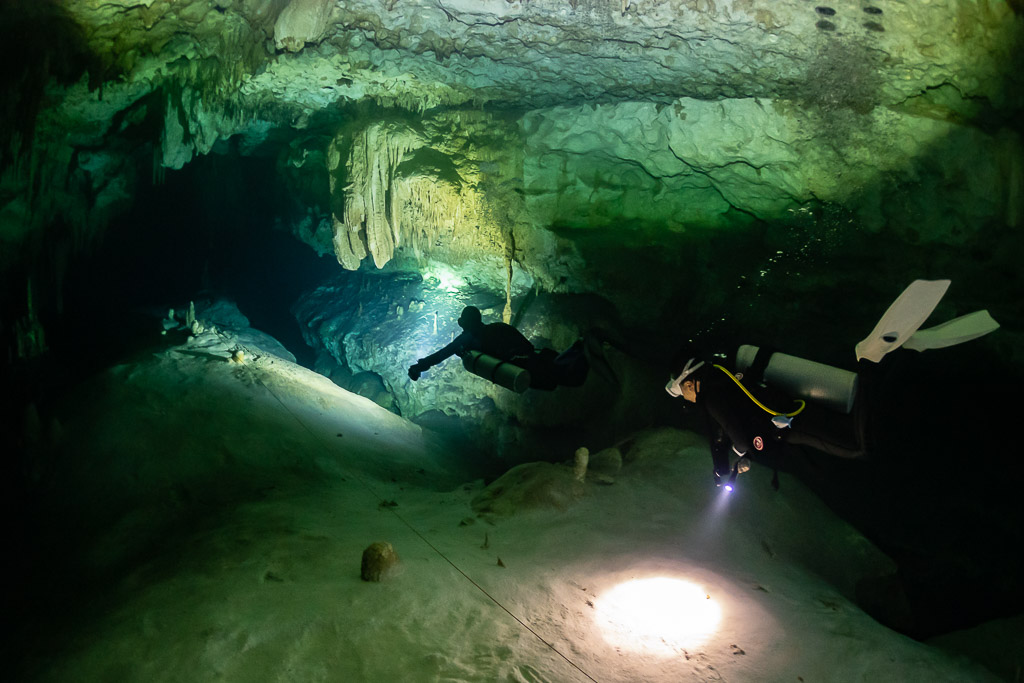 Two divers on a wide sandy cave floor, torch spot in the foreground, Quintana Roo, Mexico