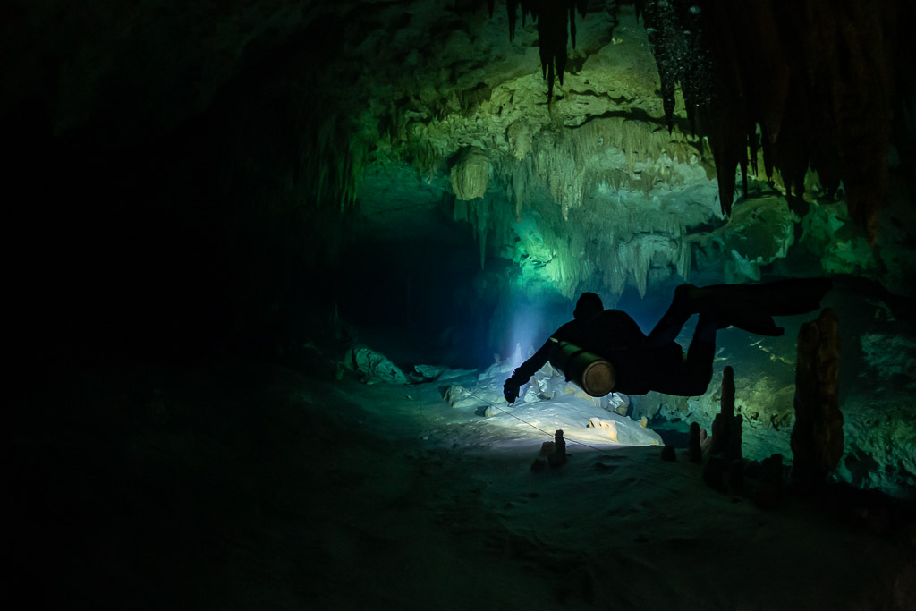 Sidemount diver in silhouette among small stalagmites, green glow behind, Quintana Roo, Mexico