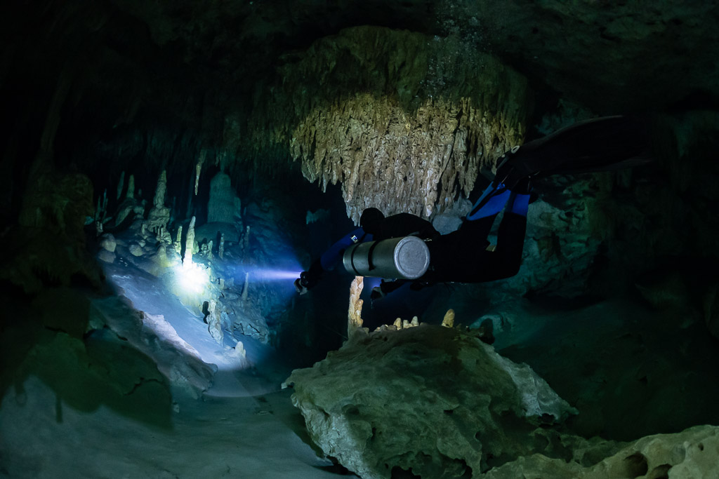 Sidemount diver with blue fins lighting a passage under a stalactite cluster, Quintana Roo, Mexico