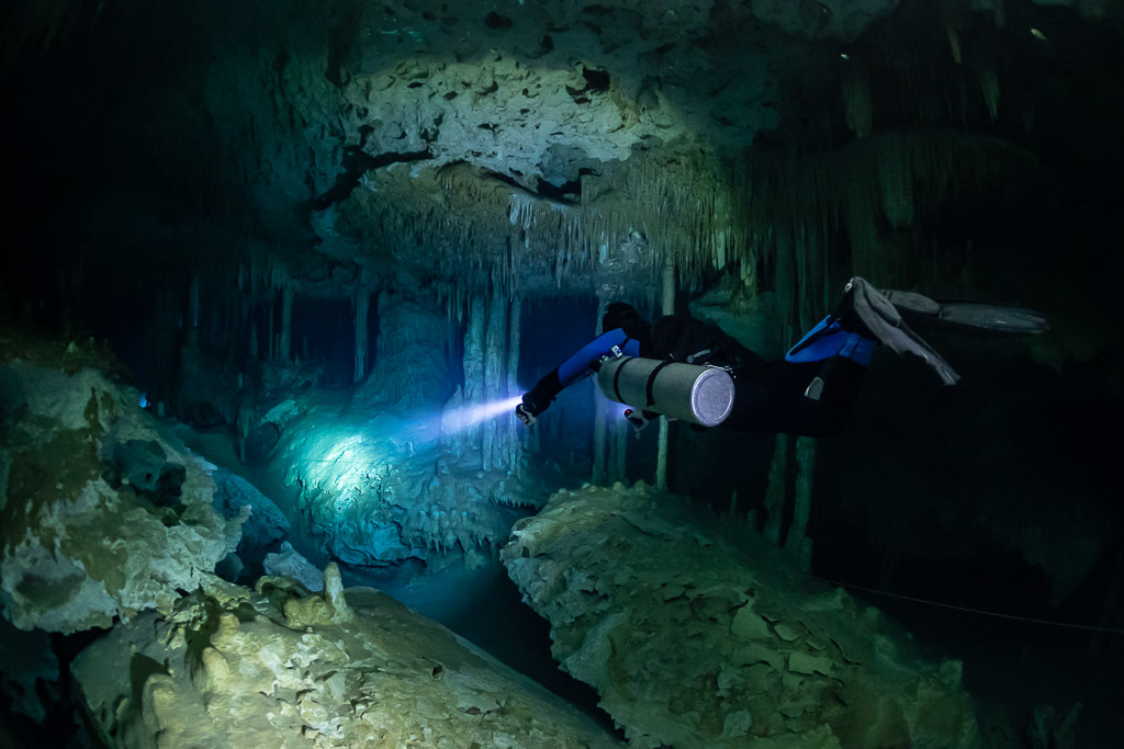 Sidemount diver lighting a green cave room above pale boulders, Quintana Roo, Mexico