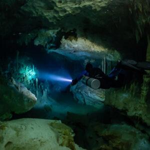 Sidemount diver passing over boulders into a green-lit room, Quintana Roo, Mexico