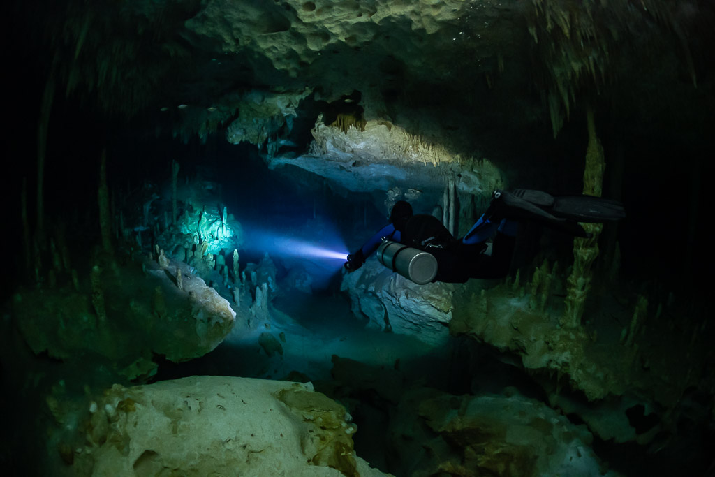 Sidemount diver passing over boulders into a green-lit room, Quintana Roo, Mexico