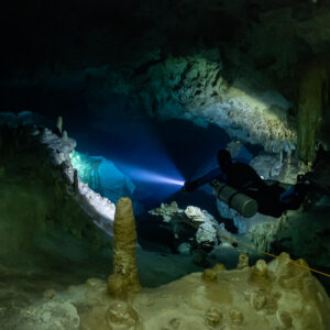 Sidemount diver beaming past a stalagmite toward a pale passage, Quintana Roo, Mexico