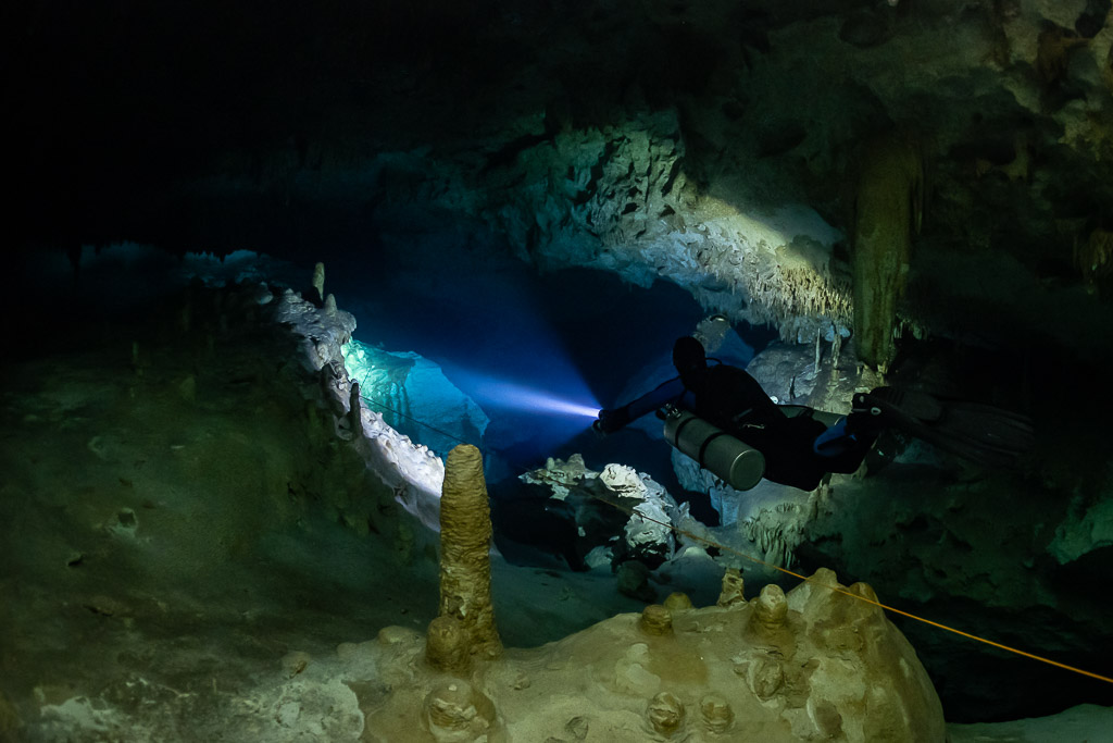 Sidemount diver beaming past a stalagmite toward a pale passage, Quintana Roo, Mexico