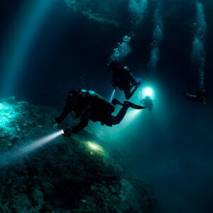 Divers crossing sunbeams above a rock mound with bubble trails, Quintana Roo, Mexico