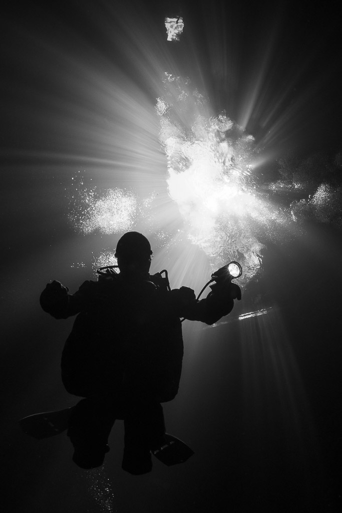 Silhouette de plongeur vue d’en dessous devant l’ouverture lumineuse d’un cénote, en noir et blanc, Quintana Roo, Mexique