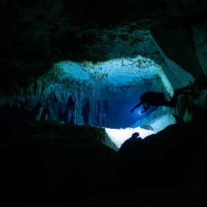 Cave diver passing a low turquoise-lit opening between dark rock walls, Quintana Roo, Mexico
