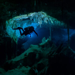 Cave diver silhouetted under a stalactite-fringed arch in blue water, Quintana Roo, Mexico