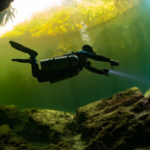 Sidemount diver gliding under a cavern arch toward green daylight, Casa Cenote, Quintana Roo, Mexico
