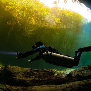 Sidemount diver framed by a rocky arch with yellow-green mangrove light, Casa Cenote, Quintana Roo, Mexico
