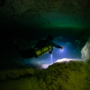 Sidemount diver lighting a rock in a dark green cavern, Casa Cenote, Quintana Roo, Mexico