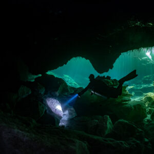 Sidemount diver passing a cavern opening with turquoise light beyond, Casa Cenote, Quintana Roo, Mexico