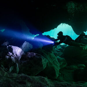 Sidemount diver with torch gliding out of a cavern into turquoise light, Casa Cenote, Quintana Roo, Mexico