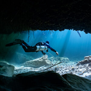 Sidemount diver exiting a dark cavern toward blue sunlit water, Casa Cenote, Quintana Roo, Mexico