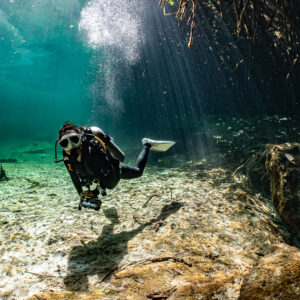 Diver approaching over clear sand beside a dark boulder, Casa Cenote, Quintana Roo, Mexico