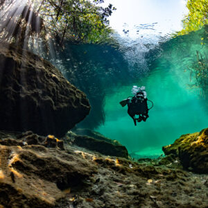 Diver in a turquoise channel between rocks under backlit sun rays, Casa Cenote, Quintana Roo, Mexico