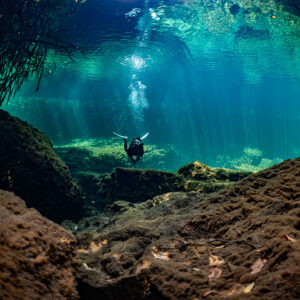 Diver framed by brown boulders in a turquoise cenote basin, Casa Cenote, Quintana Roo, Mexico