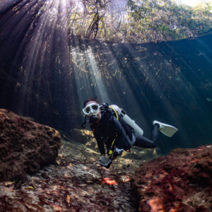 Woman diver in a Santa hat between reddish rocks under sun rays, Casa Cenote, Quintana Roo, Mexico
