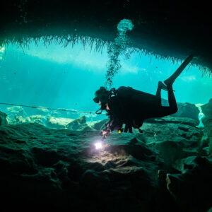 Diver with torch under a cavern arch facing turquoise daylight, Casa Cenote, Quintana Roo, Mexico