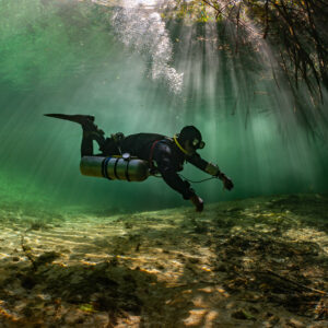 Sidemount diver gliding over sand under mangrove roots and sunrays, Casa Cenote, Quintana Roo, Mexico