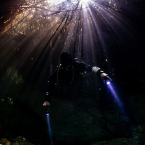 Diver with torches in dark cavern below a starburst of daylight, Casa Cenote, Quintana Roo, Mexico