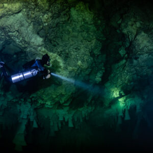 Sidemount diver lighting a rough limestone wall in dark green water, Cenote El Zapote, Quintana Roo, Mexico