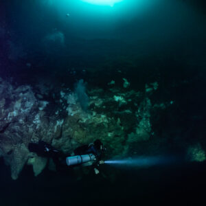 Small sidemount diver lighting the wall under a distant glowing opening, Cenote El Zapote, Quintana Roo, Mexico