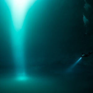 Tiny diver with torch beside a vast shaft of turquoise light, Quintana Roo, Mexico