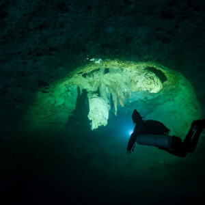 Sidemount diver silhouetted against a green-lit cave with stalactites, Cenote El Zapote, Quintana Roo, Mexico