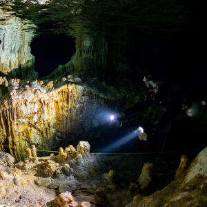 Distant divers beaming torches past a golden flowstone wall, darker frame, Quintana Roo, Mexico
