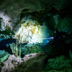 Sidemount diver lighting a pale rock face in a green cavern, Quintana Roo, Mexico