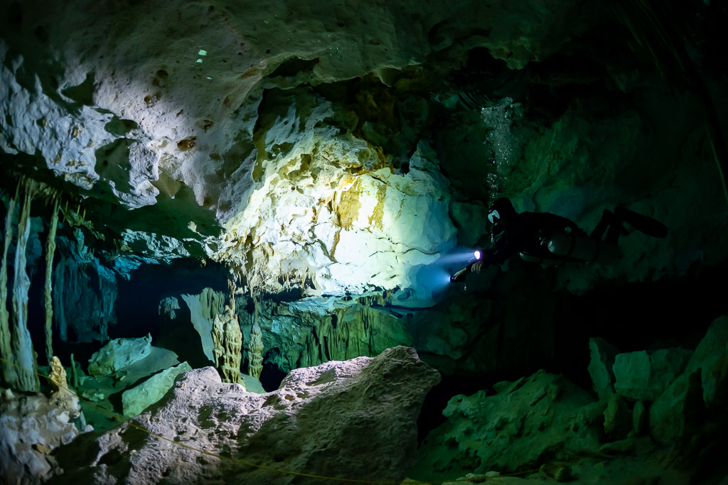 Plongeur en sidemount éclairant un pan rocheux clair dans une caverne verte, Quintana Roo, Mexique
