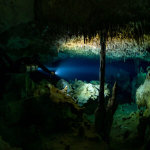 Diver beaming torch under a low ceiling beside a slender column, Quintana Roo, Mexico