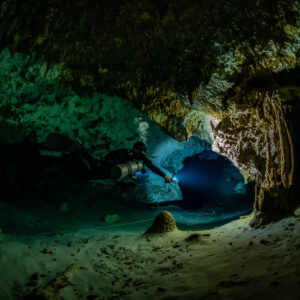 Sidemount diver lighting a low dark passage over a sandy floor, Quintana Roo, Mexico