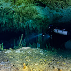 Sidemount diver above a lit pale floor under hanging stalactites, Quintana Roo, Mexico