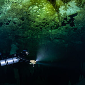 Sidemount diver beaming into darkness under a pitted green ceiling, Quintana Roo, Mexico