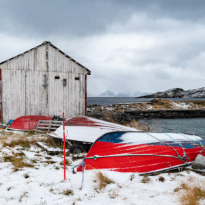 Weathered wooden boathouse with red rowing boats upturned in the snow, Seglvik, Troms, Norway