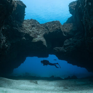 Diver silhouetted under a rock arch above white sand, Flic-en-Flac, Mauritius