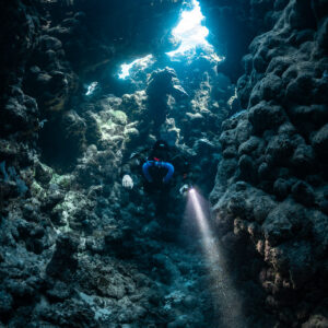 Rebreather diver with torch in a narrow cave shaft lit from above, Zabargad, Red Sea, Egypt