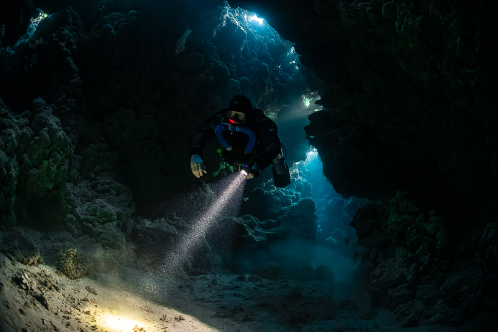 Rebreather diver emerging from the cave darkness with torch, Zabargad, Red Sea, Egypt