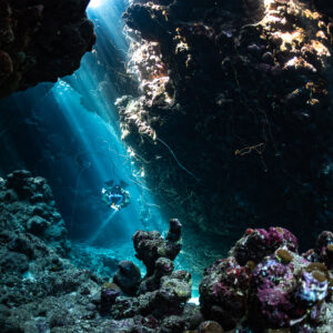 Distant rebreather diver in a cave lit by sunbeams, coral heads in front, Gota Jeny, Red Sea, Egypt