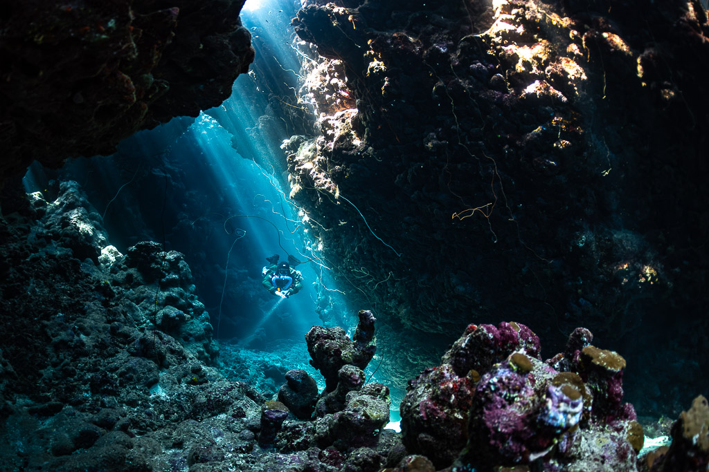 Distant rebreather diver in a cave lit by sunbeams, coral heads in front, Gota Jeny, Red Sea, Egypt