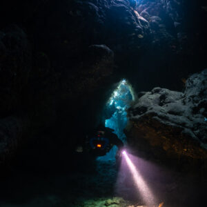 Rebreather diver passing through a cave doorway, torch beam on the floor, Saint John's Cave, Red Sea, Egypt