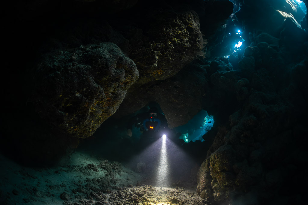 Rebreather diver emerging between boulders in the dark cave, Saint John's Cave, Red Sea, Egypt