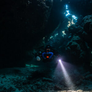 Rebreather diver below a line of skylights, torch beam angled down, Saint John's Cave, Red Sea, Egypt
