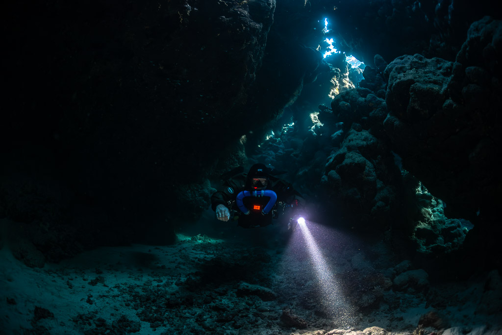 Rebreather diver below a line of skylights, torch beam angled down, Saint John's Cave, Red Sea, Egypt