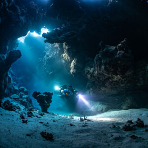 Distant rebreather diver in a vast sandy cave hall with sunbeams, Saint John's Cave, Red Sea, Egypt