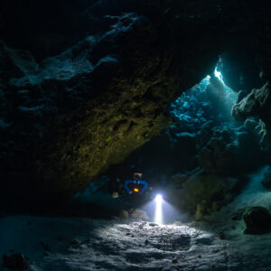 Rebreather diver under a huge cave arch, torch lighting the sand, Saint John's Cave, Red Sea, Egypt