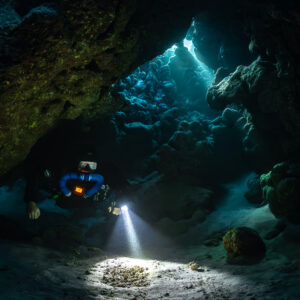 Rebreather diver under a low cave ceiling, torch on the sand, Saint John's Cave, Red Sea, Egypt